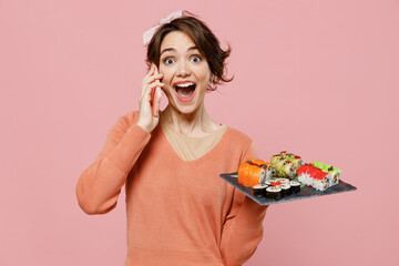 Wall Mural - Young surprised amazed shocked woman in sweater hold in hand makizushi sushi roll served on black plate traditional japanese food talk on mobile cell phone isolated on plain pastel pink background