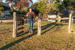 Caucasian man person walking through an open gate in a Texas field wire fence through a grassy field with trees, a well house and water tank in the background..