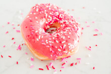 A Donut With Pink Frosting And Valentines Pink, White, And Red Heart Sprinkles On A White Marble Background