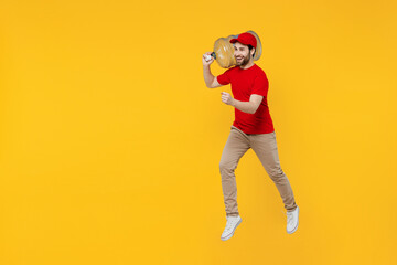 Wall Mural - Professional side view fun delivery guy employee man in red cap T-shirt uniform workwear work as dealer courier hold water bottle jump high isolated on plain yellow background studio. Service concept.