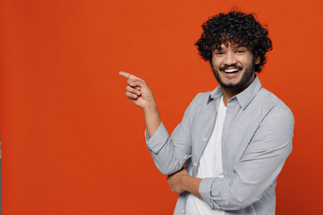 Poster - Smiling fascinating happy young bearded Indian man 20s years old wears blue shirt pointing index finger aside on workspace area copy space mock up isolated on plain orange background studio portrait.