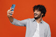 © ViDi Studio - Fun fascinating friendly young bearded Indian man 20s years old wear blue shirt doing selfie shot on mobile cell phone post photo on social network isolated on plain orange background studio portrait