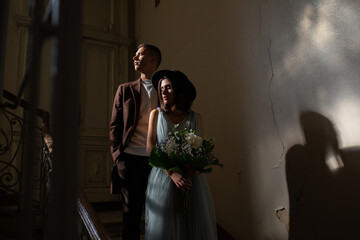 Wall Mural - Newlyweds in love stand in an old dark building with shadows. Portrait of a stylish groom with coat and a beautiful cute bride in a blue dress and black hat.