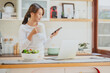 © interstid - Smiling beautiful asian woman using smartphone mobile in the kitchen while drinking coffee in morning