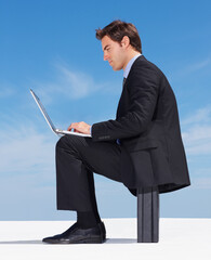 Young executive working on a laptop against clear sky. Side view of a successful young business man working on a laptop against clear sky.