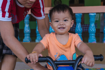 Little boy learning to ride a bicycle at home with sister or mother. Asian mother teaches her son to play baby balance bike. Pretty young woman helping her adorable son ride a bike. Family concept