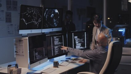 Medium long of young Asian female manager talking to Caucasian male programmer, who sitting at desk, using computer in corporate office at night, Black man talking on phone on background