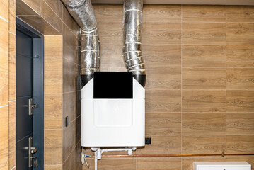 Home mechanical ventilation with heat recovery hanging on the wall in a modern gas boiler room with brown ceramic tiles imitating wood.