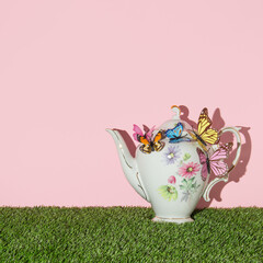 Wall Mural - Spring creative layout with english garden teapot with colorful butterflies on green grass and pastel pink background. 80s, 90s retro romantic aesthetic summer concept. Minimal  fashion tea idea.