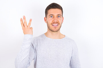 young caucasian man wearing grey sweater over white background smiling and looking friendly, showing number three or third with hand forward, counting down