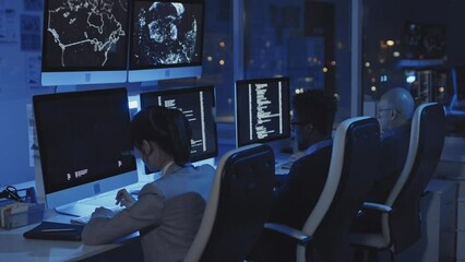 Side-view of three diverse programmers sitting on chairs at desks in dark modern office, working on computers at night