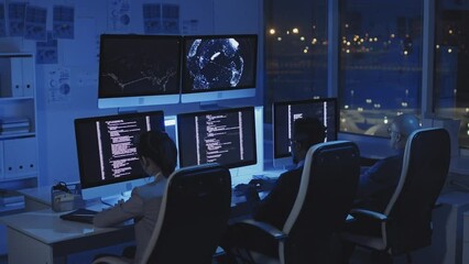 Rear of three multiethnic programmers sitting on chairs at desks in dark modern office, coding on computers at night