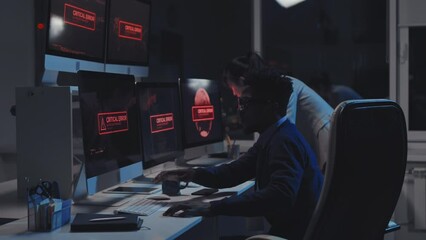 Side-view of two young diverse cyber security specialists fixing critical error, warning alarm showing on multiple computers monitors on desk in office at night