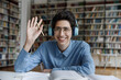© fizkes - Happy young Jewish male student in eyeglasses wearing headphones making hello gesture looking at camera, starting video call conversation with teacher, sitting at table in library, e-learning concept.