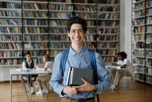 Portrait Of Smiling Millennial Jewish Male Student In Eyeglasses Holing Books In Hands, Posing In Modern College Library. Joyful Handsome Young Guy With Backpack Looking At Camera, Education Concept.