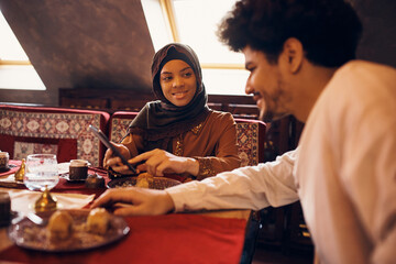 Wall Mural - Happy Muslim couple uses cell phone while drinking coffee and having dessert at home.