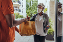 Woman With Protective Face Mask Delivering Food Bag To Man At Doorway