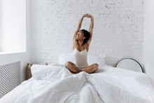 Woman Sitting Cross-legged With Arms Raised On Bed At Home