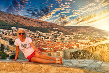Girl Sitting On Top Of Fort Lovrijenac Fortress, Over The West Harbour With Fort Bokar. Dubrovnik City Walls In Croatia At Sunset. Dubrovnik Historic City In Dalmatia And UNESCO World Heritage Site.