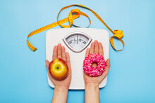 Diet Concept. Female Hands Hold Sweet Donut And Red Apple Against The Background Of Scales And A Measuring Tape, Choice Between Healthy Unhealthy Food Dessert. Top View