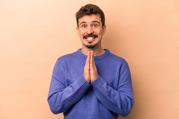 Wall Mural - Young caucasian man isolated on beige background holding hands in pray near mouth, feels confident.