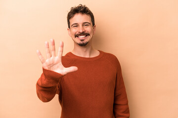 Wall Mural - Young caucasian man isolated on beige background smiling cheerful showing number five with fingers.