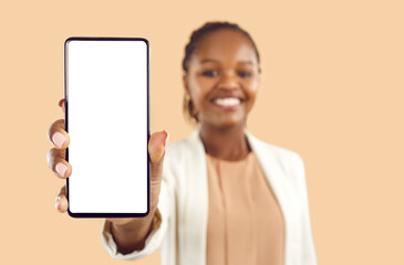 Smiling African American woman isolated on orange studio background show white mockup cellphone screen. Happy black female demonstrate deal or sale on phone. Focus on smartphone, blurred background.