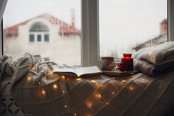 Sticker - Cup of hot drink, open book and Christmas lights on knitted blanket near window. Cozy season