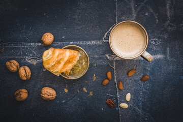 Wall Mural - Piece of delicious French croissant dipped in jam, a cup of coffee and some nuts, breakfast scene, black background, top view