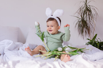 baby in a green bodysuit with white rabbit ears is sitting on a bed with a bouquet of white tulips