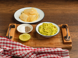 Wall Mural - A bowl with fresh guacamole on a wooden tray and potatoes chips on a white plate over a wooden table