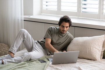 Aufkleber - Young businessman in casualwear searching through online information in laptop while lying on comfortable bed in bedroom