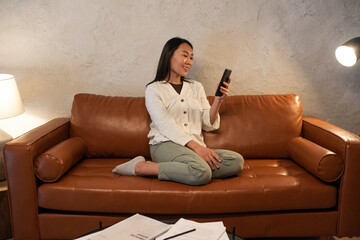 Aufkleber - Young smiling Asian woman communicating in video chat while sitting on sofa and holding smartphone in front of herself