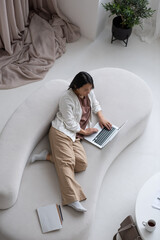 Aufkleber - Young serious Asian female employee in casual clothes lying on sofa while networking or doing online shopping at home