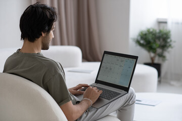 Aufkleber - Young contemporary man with laptop on knees looking through working plan on screen while sitting on sofa in living room