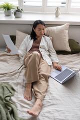 Aufkleber - Young Asian female auditor with papers sitting on double bed and looking through online data about business development