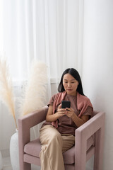 Aufkleber - Young serious Asian woman in casualwear texting in mobile phone while sitting in armchair by window covered with chiffon curtains