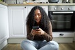 © Anatoliy Karlyuk - Pretty African American young woman in casual brown sweater and light-blue jeans sitting on floor with legs folded, chatting with friends on her phone, online, before cooking lunch, smiling