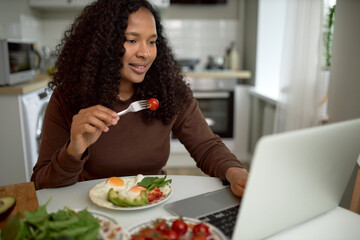 Woman having breakfast and watching favorite series on laptop, sitting at kitchen table with cherry tomato on her fork and rich tasty breakfast in her plate with eggs and vegetables