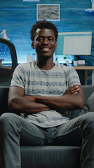 Poster - African american adult sitting on couch and looking at camera. Portrait of black person with crossed hands smiling while being at home with technology. Man of african ethnicity