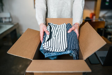 Business Woman prepares a package in a cardboard box for shipping with clothes from her online store - Millennial sells second-hand used clothing in her home - Start up concept