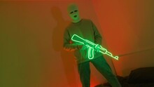 A Gangster In A White Mask Holds A Neon Machine Gun. A Young Guy Is Standing In An Ultraviolet Room On A Bed. Video 4k For A Rap Clip In A Neon Room.