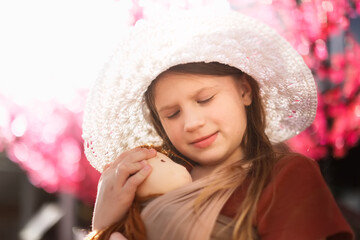 Wall Mural - Cute long-haired european kid girl with doll in sling, spring photography. Girl with doll on background of flowering trees. conscious natural parenting, doll play