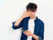 Portrait of handsome Chinese young man with black curly hair in blue shirt posing against white wall background. Fingers rubbing eyes with mobile phone in hand, looks tired, front view studio shot.