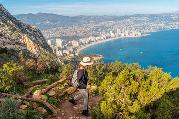 Wall Mural - A young hiker wearing a hat on the descent path of the Penon de Ifach Natural Park with the city of Calpe in the background, Valencia. Spain. Mediterranean sea. View of La Fossa beach