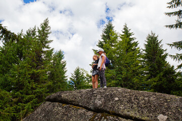 Wall Mural - Two little girl on the top of big stone in the forest. Adventure in nature. Summer day. Happy children. Green forest and blue sky with clouds in background.