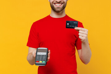 Wall Mural - Cropped delivery boy employee man in red cap T-shirt uniform workwear work as dealer courier hold wireless bank payment terminal process acquire credit card payment isolated on plain yellow background