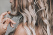 © Марина Демешко - A young blonde woman with the wavy hair holding a lock of hair in her hand isolated on a gray background. Result of coloring, highlighting, perming. Beauty and fashion