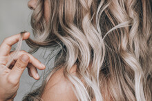 A Young Blonde Woman With The Wavy Hair Holding A Lock Of Hair In Her Hand Isolated On A Gray Background. Result Of Coloring, Highlighting, Perming. Beauty And Fashion