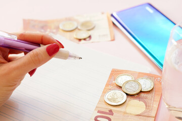 Sticker - Closeup shot of a woman holding a pen, money, notebook, water and a smartphone on a pink background